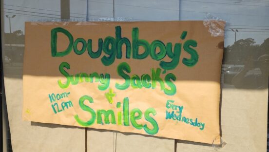 This photo shows a handwritten, makeshift sign promoting Doughboy's Sunny Sacks & Smiles program on the pizzeria's front window.