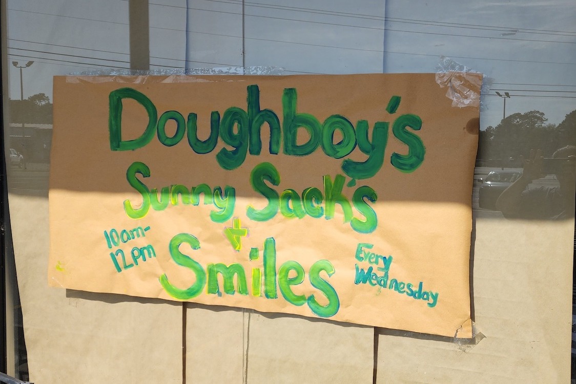 This photo shows a handwritten, makeshift sign promoting Doughboy's Sunny Sacks & Smiles program on the pizzeria's front window.