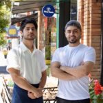 This photo shows the two brothers, one wearing a white button-down shirt and the other in a white T-shirt, standing outside in front of their pizzeria.