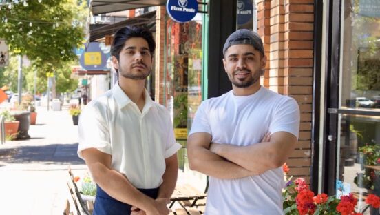 This photo shows the two brothers, one wearing a white button-down shirt and the other in a white T-shirt, standing outside in front of their pizzeria.