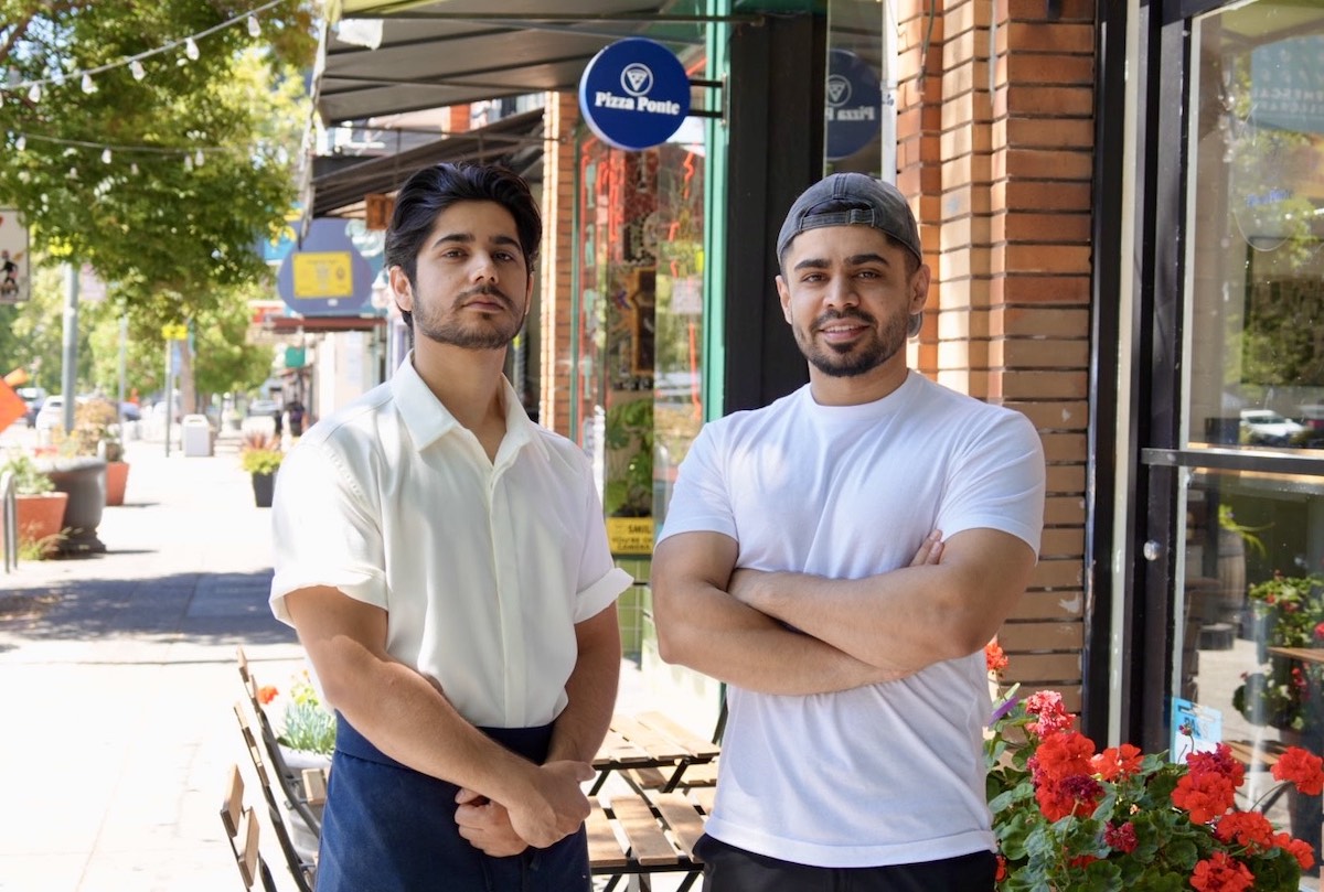 This photo shows the two brothers, one wearing a white button-down shirt and the other in a white T-shirt, standing outside in front of their pizzeria.
