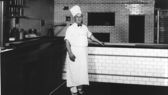 This vintage black-and-white photo shows a younger Frank Pepe in his pizzeria's kitchen, wearing a tall chef hat, a black bowtie, a white shirt and pants and a white apron.