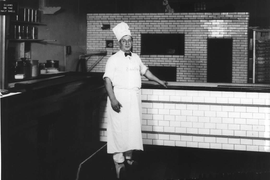 This vintage black-and-white photo shows a younger Frank Pepe in his pizzeria's kitchen, wearing a tall chef hat, a black bowtie, a white shirt and pants and a white apron.