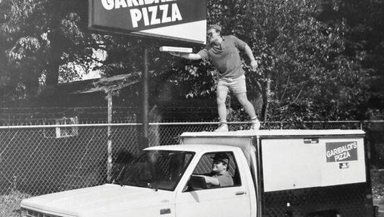 This photo shows a man standing on top of a white truck, holding out a pizza box underneath a sign that says Garibaldi's Pizza.
