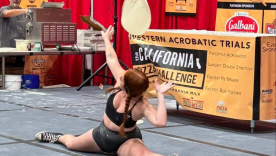 This photo shows a young woman with long braided here doing a split while tossing dough over her shoulder at the California Pizza Challenge.