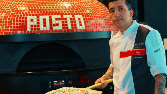 This photo shows Juan Perez, wearing a white 50 Top Pizza chef coat, standing in front of his restaurant's pizza oven with an unbaked pizza on a wooden peel.