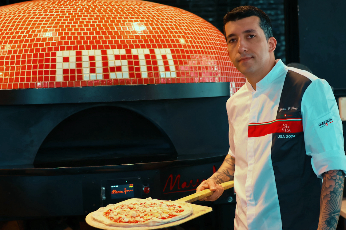This photo shows Juan Perez, wearing a white 50 Top Pizza chef coat, standing in front of his restaurant's pizza oven with an unbaked pizza on a wooden peel.