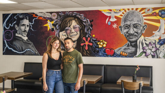 This photo shows Maggie and Giovanni Canepa, arms around each other, standing in front of a colorful mural featuring Nikola Tesla, Janis Joplin and Mahatma Gandhi.