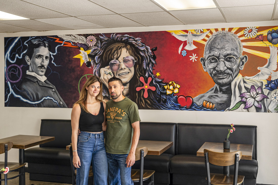 This photo shows Maggie and Giovanni Canepa, arms around each other, standing in front of a colorful mural featuring Nikola Tesla, Janis Joplin and Mahatma Gandhi.