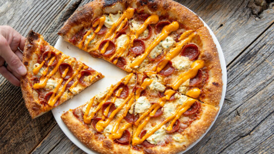 This photo shows a slice being removed from a pizza topped with pepperoni, ricotta and a drizzle of something brownish.