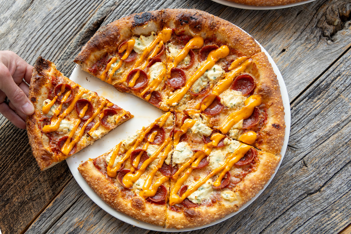 This photo shows a slice being removed from a pizza topped with pepperoni, ricotta and a drizzle of something brownish.