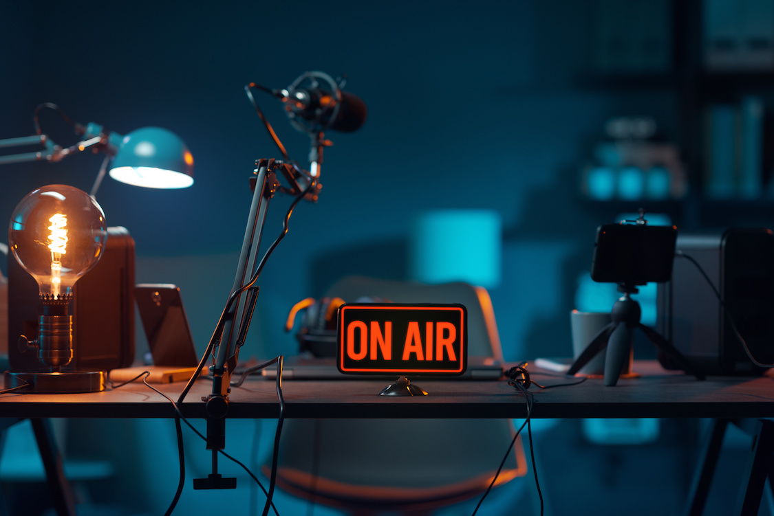This photo shows a podcast studio desk with microphones and a sign that says "On Air" in red letters.