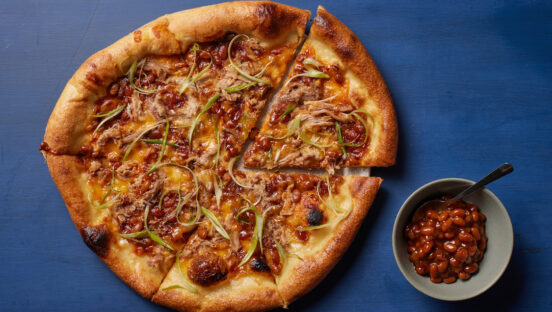 This photo shows a pizza topped with pork and beans set against a blue background with a small bowl of beans next to it.