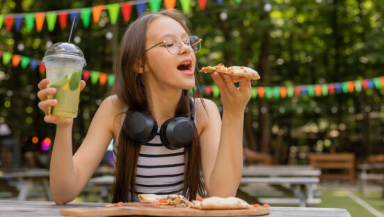 This photo shows a young woman, possibly a teenager, getting ready to take a bite of a pizza slice with one hand, a beverage in the other, and looking happy to be dining alone.