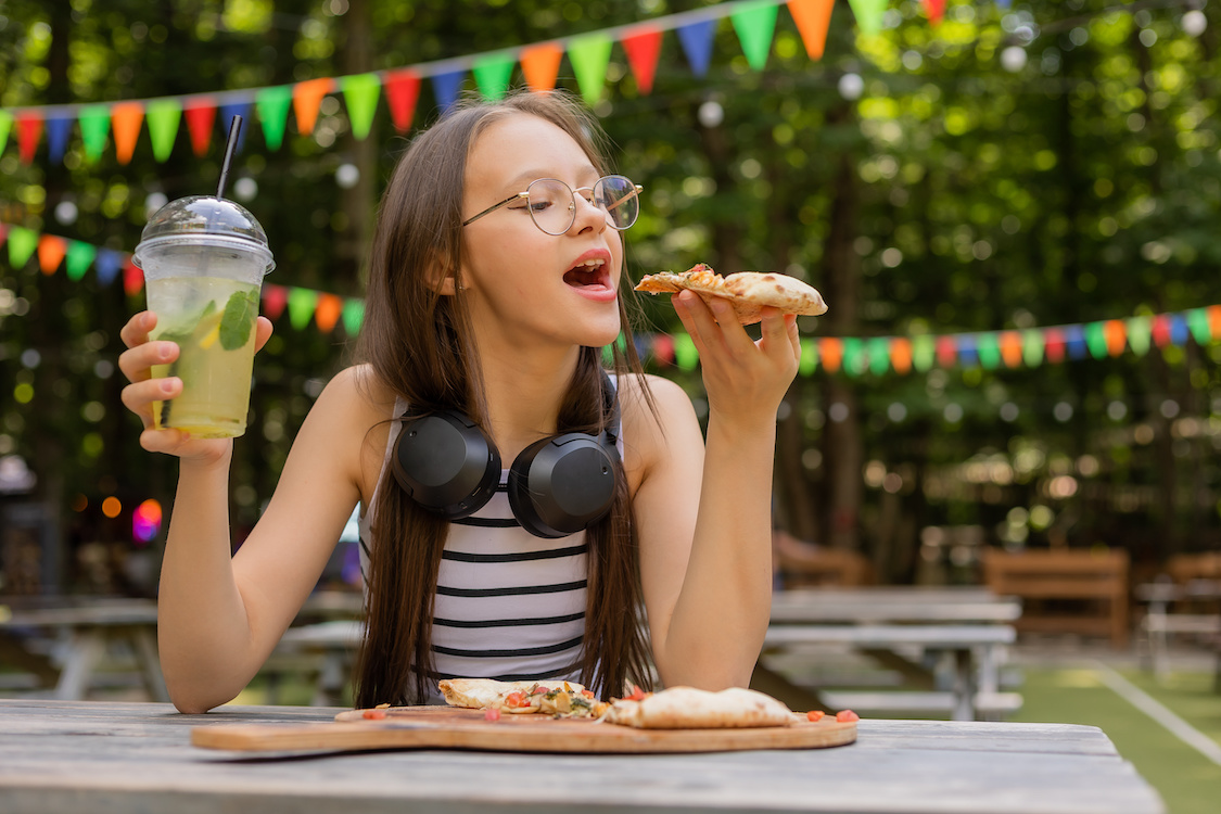 This photo shows a young woman, possibly a teenager, getting ready to take a bite of a pizza slice with one hand, a beverage in the other, and looking happy to be dining alone.