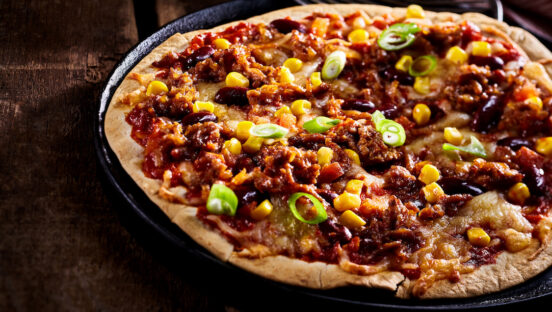 This photo shows a large flat tortilla bread pizza covered with green onions, beans and corn in cast iron pan cropped on one side
