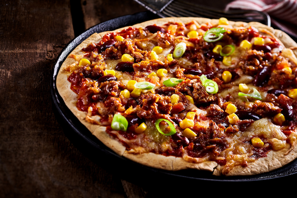 This photo shows a large flat tortilla bread pizza covered with green onions, beans and corn in cast iron pan cropped on one side