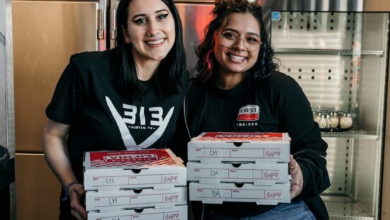 This photo shows two young female Via 313 employees standing side by side and holding pizza boxes.