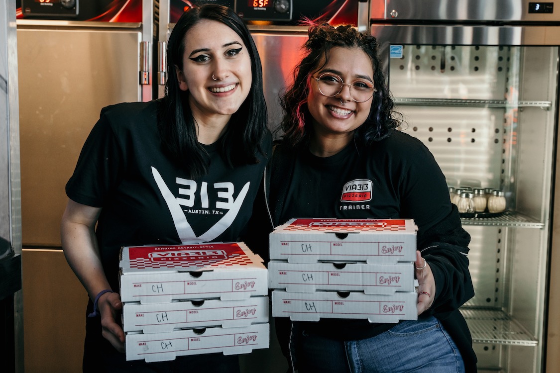 This photo shows two young female Via 313 employees standing side by side and holding pizza boxes.