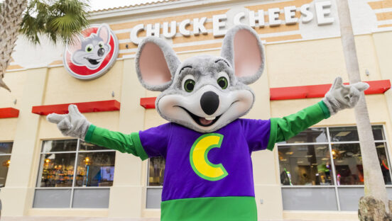 This photo shows a person in a Chuck E. Cheese costume standing in front of one the chain's restaurants with his arms spread wide open in a welcoming gesture.