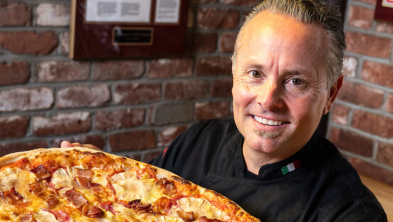 This photo shows a smiling Tony Gemignani in a black chef coat, holding one of his pizzas up to the camera.