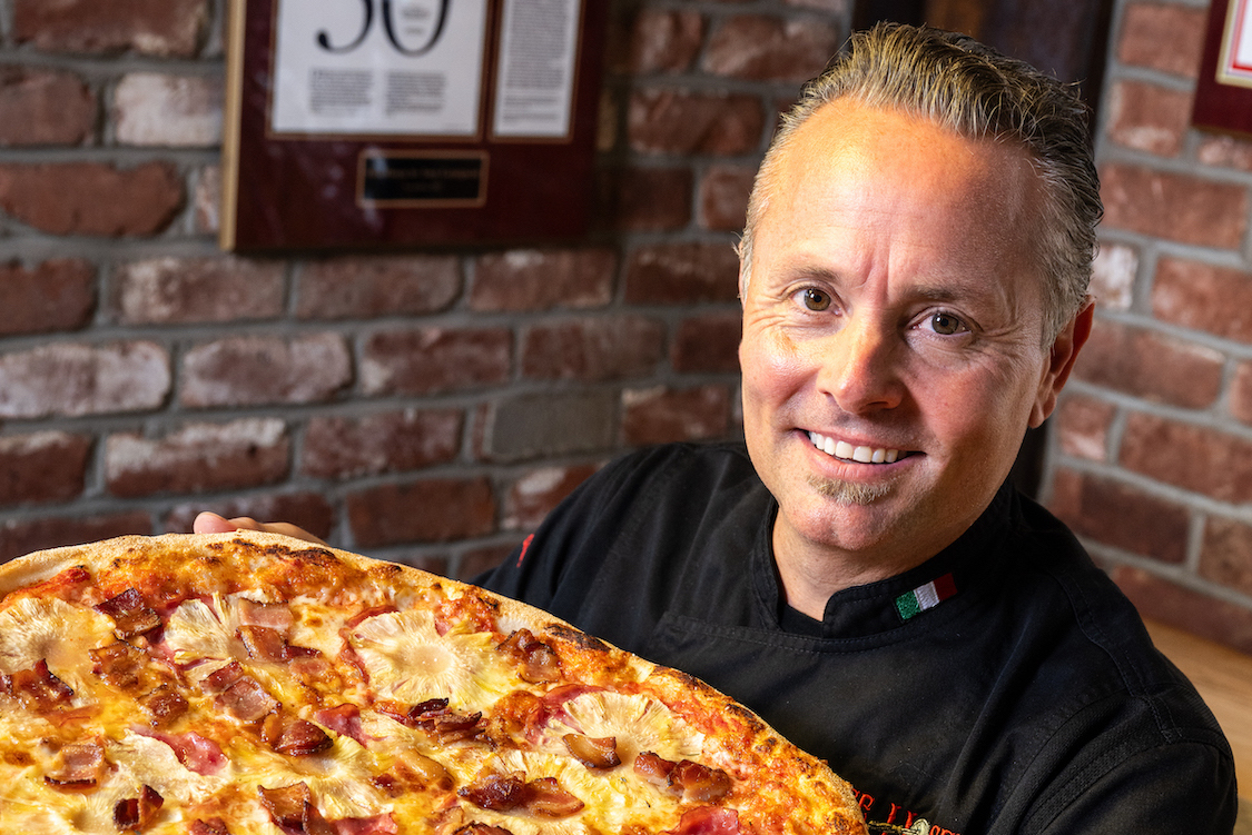 This photo shows a smiling Tony Gemignani in a black chef coat, holding one of his pizzas up to the camera.