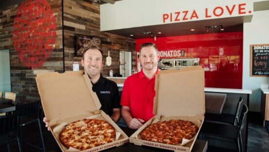 This photo shows the McLain brothers in their restaurant, holding open pizza boxes with pizzas inside.
