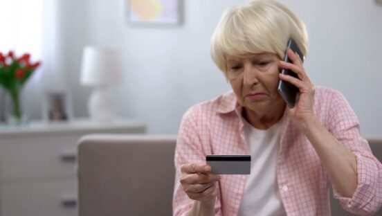 This photo shows a senior woman with a credit card talking into her phone, looking unhappy.