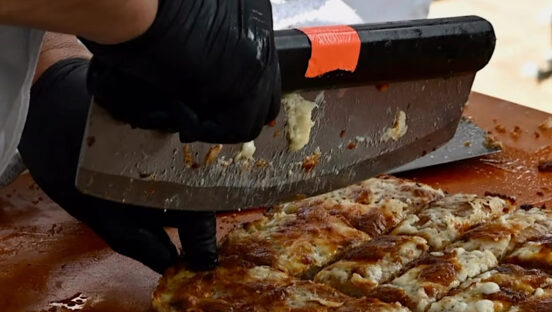 This photo shows a large slicer cutting a pepperoni and cheese pizza into rectangular slices.