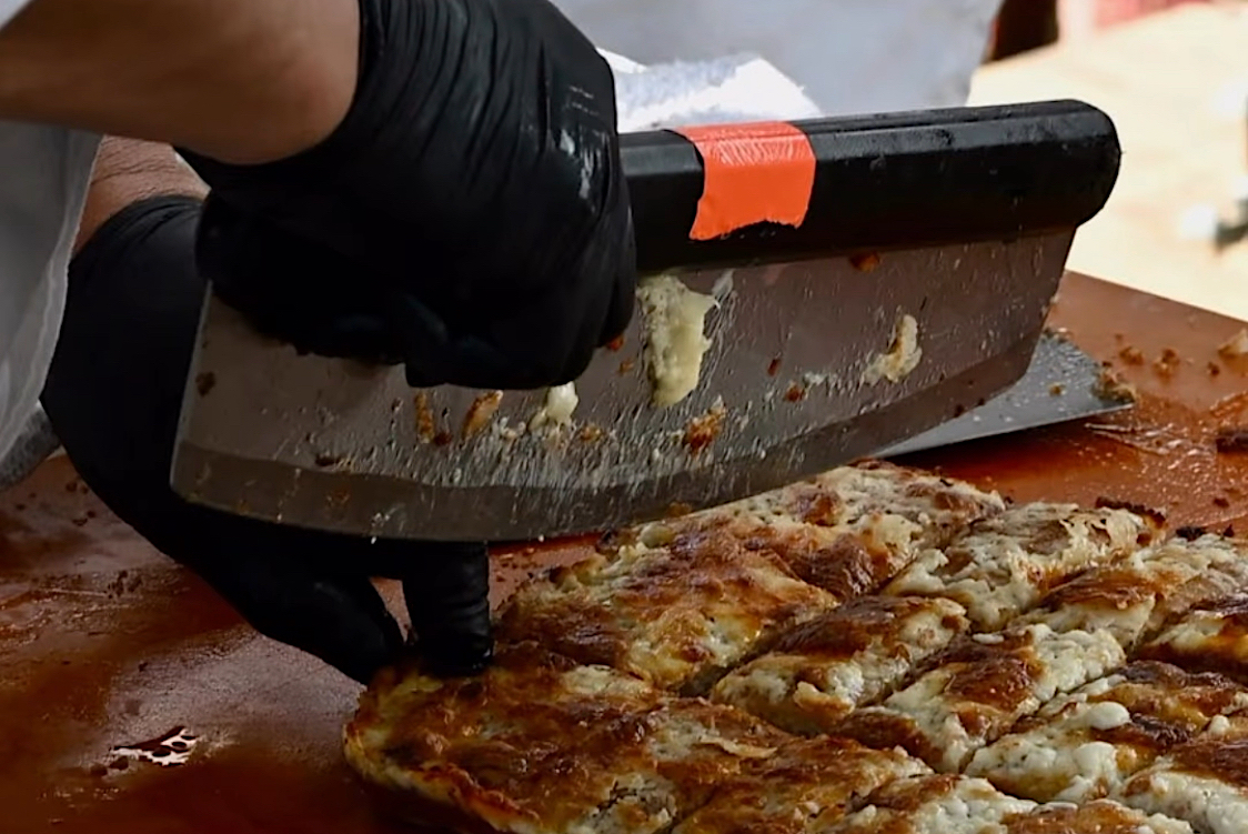 This photo shows a large slicer cutting a pepperoni and cheese pizza into rectangular slices.