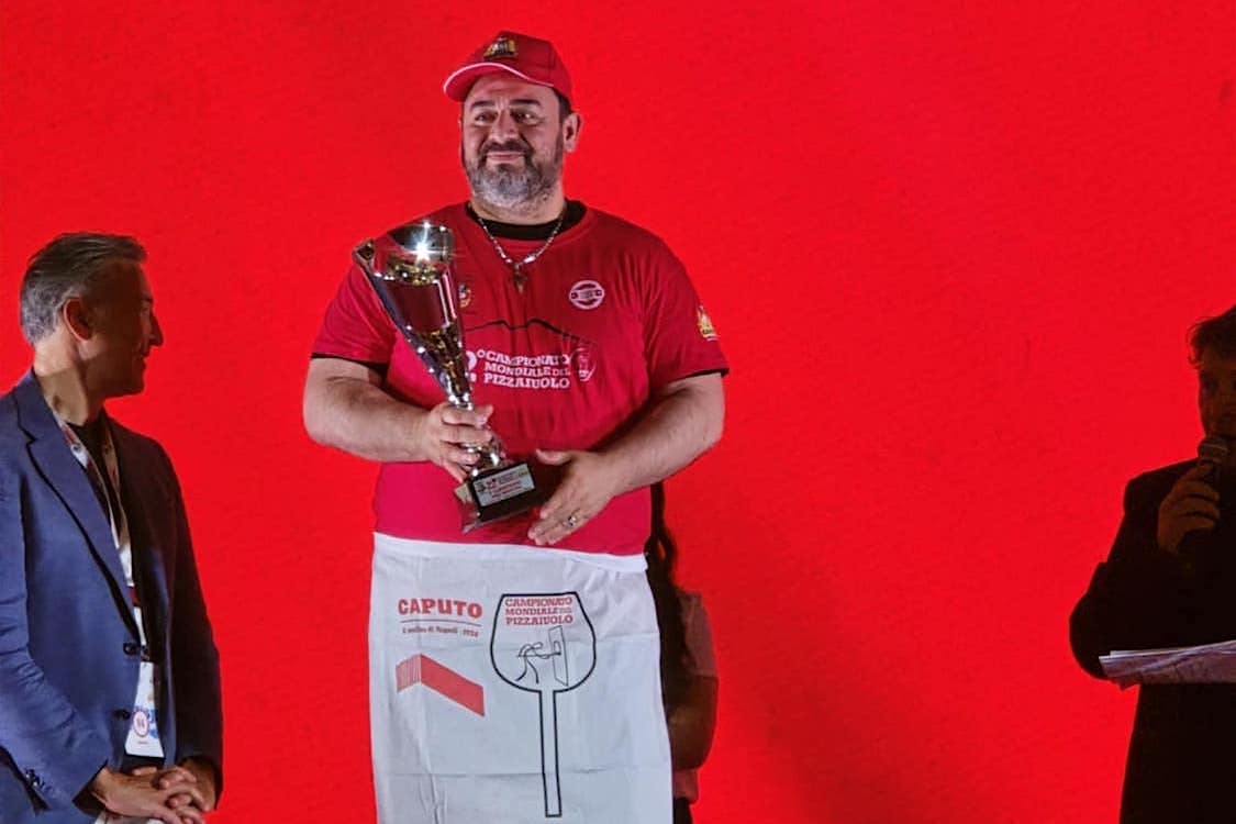 This photo shows Giovanni Labbate holding his Caputo Cup trophy and standing in front of a red backdrop.