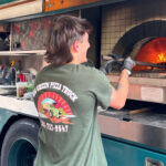 This photo shows a person in a Big Green Truck Pizza t-shirt making a pie in a wood-fired oven aboard one of the company's trucks.