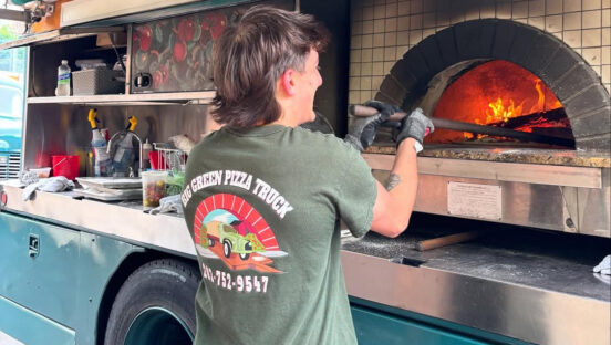 This photo shows a person in a Big Green Truck Pizza t-shirt making a pie in a wood-fired oven aboard one of the company's trucks.