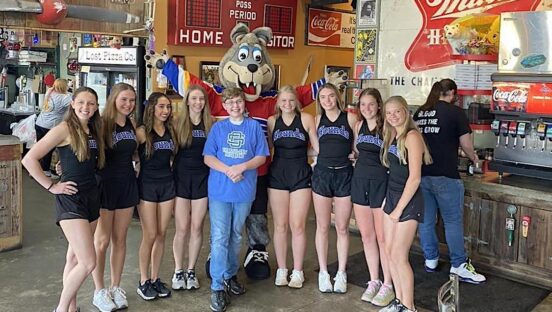 This photo shows a young boy in a blue t-shirt standing in the center of a group of teenage girls who came to Lost Pizza to sign his yearbook.