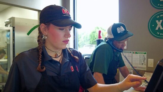 This photo shows a young female employee with pigtails tapping on the POS system screen at a Marco's Pizza restaurant.