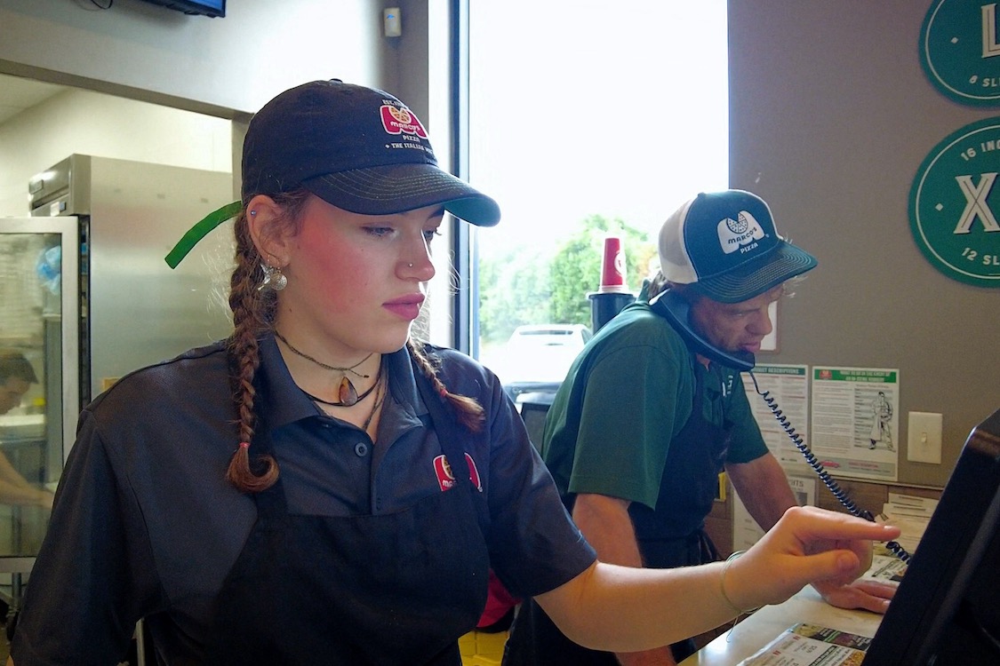 This photo shows a young female employee with pigtails tapping on the POS system screen at a Marco's Pizza restaurant.