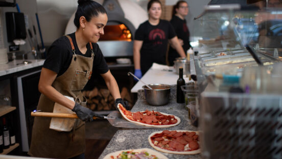 This photo shows Marisol Doyle, with her black hair in a bun and wearing a black t-shirt and brown vest, putting a pepperoni pizza on a peel while two female team members stand in the background.