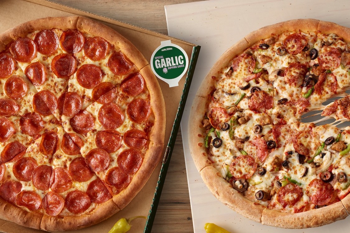 This photo shows two Papa Johns pizzas in boxes with a container of garlic sauce positioned between them.