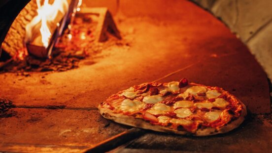 This photo shows a beautiful pizza sitting at the edge of an oven, with the wood burning further back in the photo.