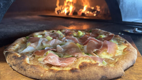 This photo shows a beautiful pizza topped with pineapple and prosciutto placed in front of a blazing wood-fired oven.