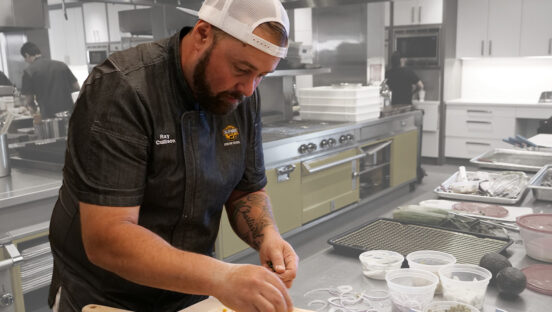 This photo shows Ray Cullison, wearing a backwards cap and a black chef coat, topping a pizza.