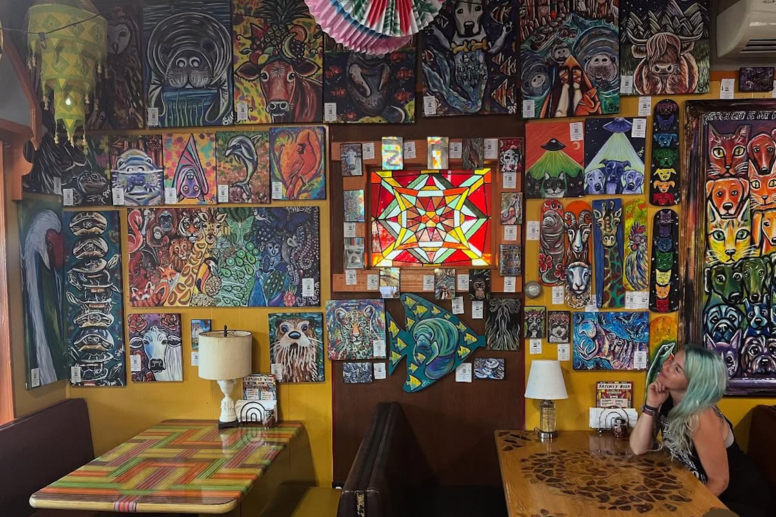 This photo shows a female customer sitting at a table and peering up at the elaborate artwork decorating one wall at Satchel's Pizza.