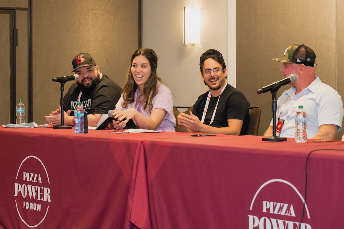 This photo shows four panelists in a dough-making session at the 2024 Pizza Power Forum. They're sitting at a table covered with a red cloth that has the Pizza Power Forum logo on it.