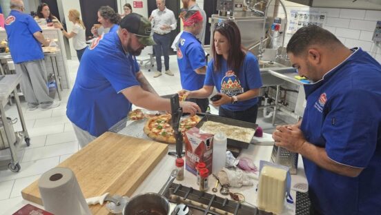This photo shows several USPT chefs gathered around a prep table as they work on making their pizzas in the Galbani kitchen.