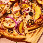This photo shows a pizza topped with peach slices, mozzarella, bacon, red onion slices and jalapeños.