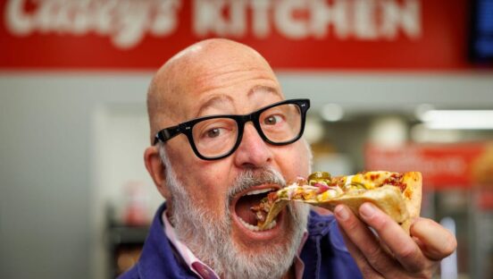 This photo shows Andrew Zimmern, with glasses and a gray beard, chomping into a slice of Casey's BBQ brisket pizza with his mouth wide open.
