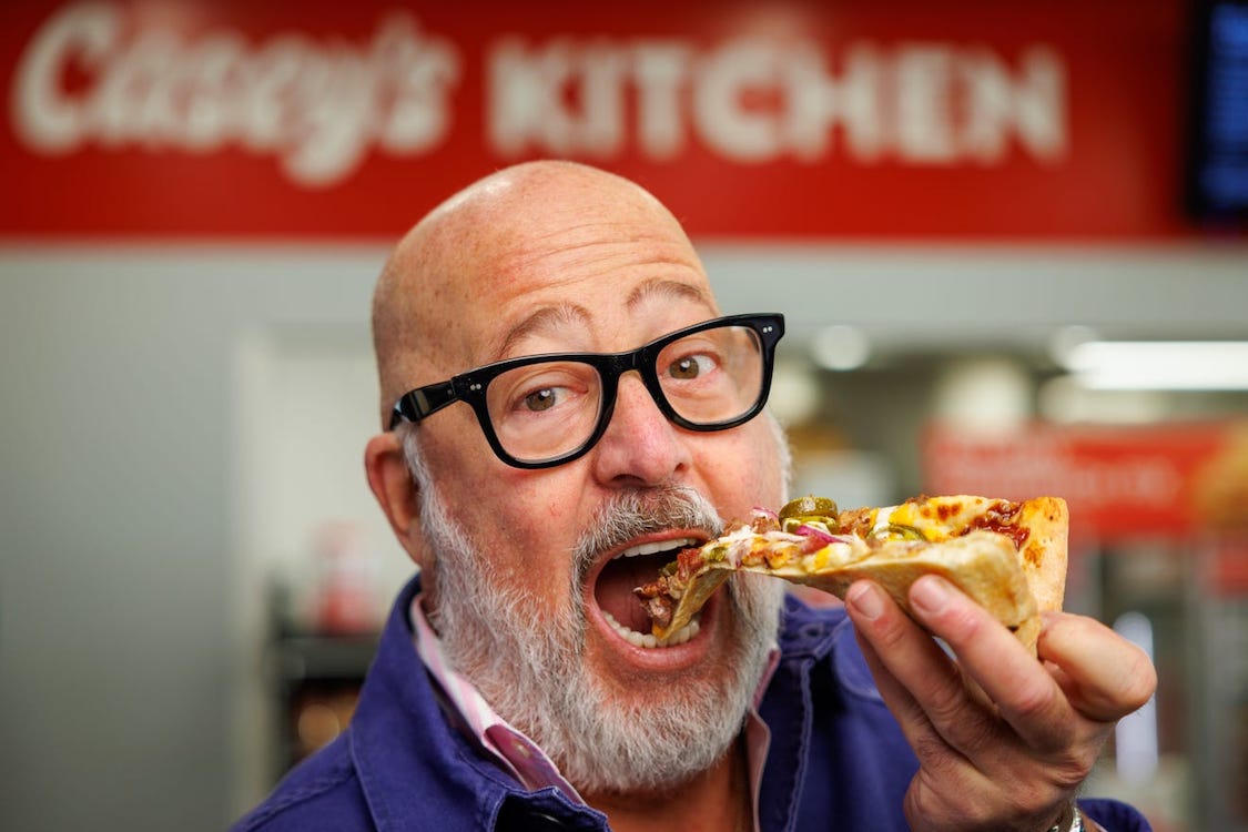 This photo shows Andrew Zimmern, with glasses and a gray beard, chomping into a slice of Casey's BBQ brisket pizza with his mouth wide open.