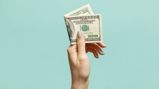This photo shows a woman's hand holding a folded $100 bill against a greenish-blue background.