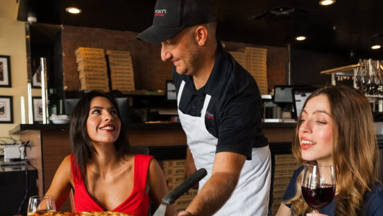 This photo shows a male server wearing a white apron and black t-shirt serving a whole pepperoni pizza to a young woman in a red dress while another woman sits next to her.