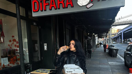 This woman shows a dark-haired woman dressed in black, eating a slice of pizza outside of Di Fara, with the restaurant's sign above her head in the background.